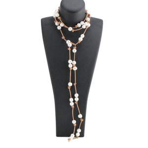 May include: A long, layered necklace featuring white pearls strung on tan leather cord. The necklace is displayed on a black mannequin, showcasing its draping design. The pearls are evenly spaced, creating a classic and elegant look.
