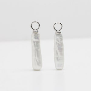 May include: Two white, elongated pearl earrings with silver-colored metal loops at the top. The pearls have a natural, irregular shape and a subtle luster, set against a plain white background.