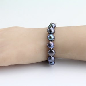 May include: A dark gray pearl bracelet. The bracelet features large, irregularly shaped pearls strung together. The pearls have a lustrous sheen, with hints of iridescent colors. The bracelet is displayed on a light-toned background.