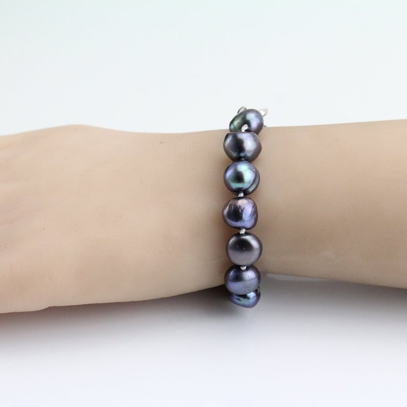 May include: A dark gray pearl bracelet. The bracelet features large, irregularly shaped pearls strung together. The pearls have a lustrous sheen, with hints of iridescent colors. The bracelet is displayed on a light-toned background.