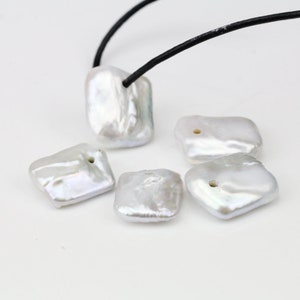 Puede incluir: Cinco perlas de agua dulce de forma cuadrada, de color blanco, con un agujero perforado en el centro. Las perlas están dispuestas sobre una superficie blanca.