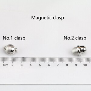 May include: Two silver magnetic clasps for jewelry making. The clasps are shown next to a ruler with measurements in centimeters. The first clasp is round and the second clasp is oval.