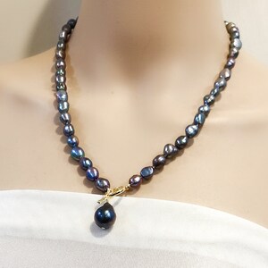Op de afbeelding: Een zwarte parelketting met een grote zwarte parel hanger. De ketting is gemaakt van kleine, onregelmatig gevormde parels en heeft een gouden sluiting.