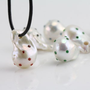 Pode incluir: Um colar com um pingente de p&eacute;rola barroca branca adornado com pedras preciosas vermelhas. A p&eacute;rola tem uma forma irregular e uma superf&iacute;cie lisa e brilhante. O colar &eacute; feito de um cord&atilde;o de couro preto.