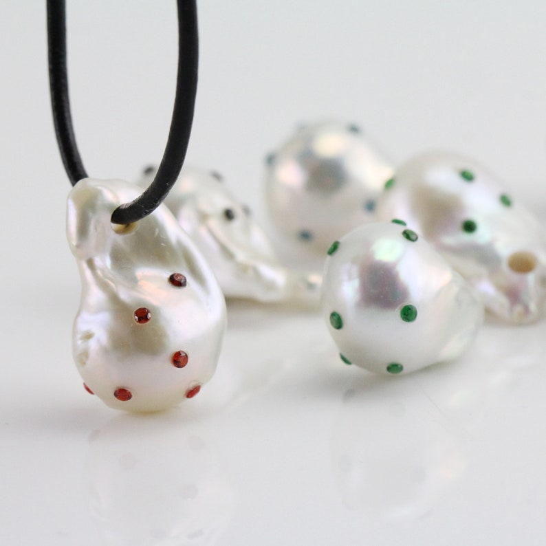 Pode incluir: Um colar com um pingente de p&eacute;rola barroca branca adornado com pedras preciosas vermelhas. A p&eacute;rola tem uma forma irregular e uma superf&iacute;cie lisa e brilhante. O colar &eacute; feito de um cord&atilde;o de couro preto.