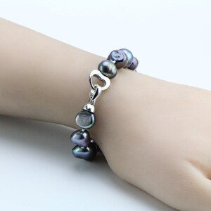 May include: A dark gray pearl bracelet with a silver heart-shaped clasp. The pearls are irregularly shaped and have a lustrous sheen. The bracelet is displayed on a light-toned background.