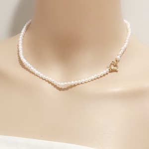 Op de afbeelding: Een delicate parelketting met een gouden hartvormige sluiting. De ketting is gemaakt van kleine, witte parels en is perfect om een ​​toets van elegantie aan elke outfit toe te voegen.
