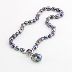 May include: A necklace made of dark grey pearls with a silver toggle clasp. The necklace features a large, irregular pearl pendant.