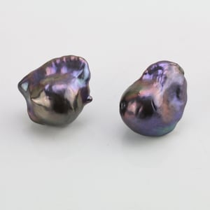 Puede incluir: Dos perlas barrocas negras iridiscentes con un tono púrpura. Las perlas tienen una forma irregular y una superficie lisa y brillante.
