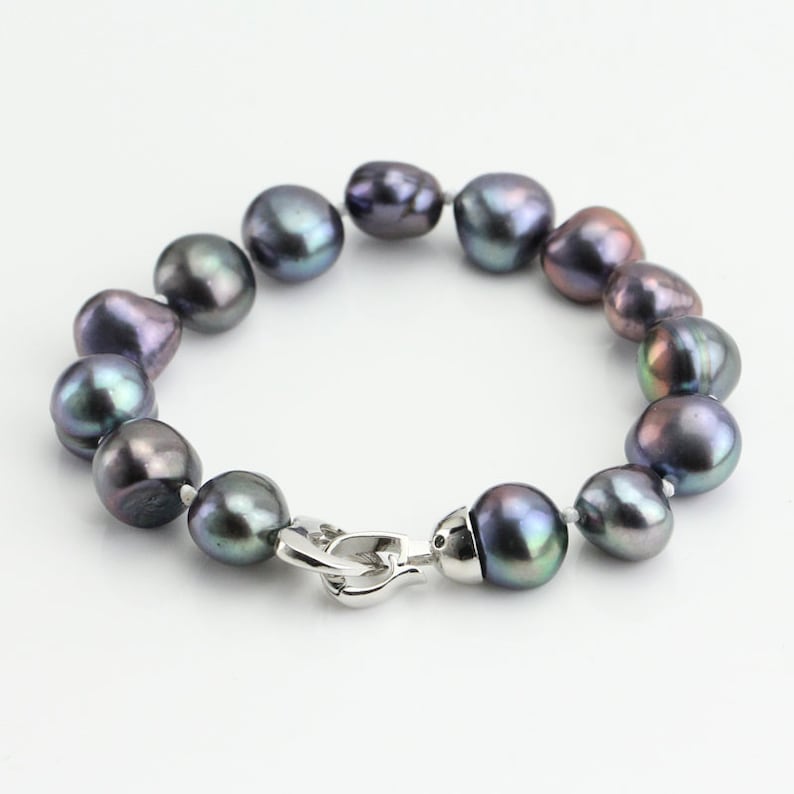 May include: A dark gray pearl bracelet with a silver clasp. The pearls are round and have iridescent hues of green, purple, and blue. The bracelet is strung on a white cord and is displayed on a white background.