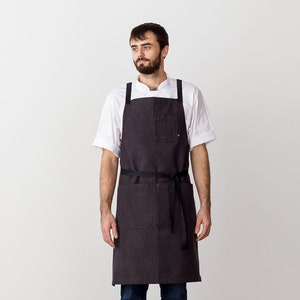 May include: A man wearing a white chef's shirt and a dark gray denim apron with a black adjustable strap. He is standing in front of a white wall.