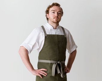 Könnte beinhalten: Ein Mann trägt eine weiße Kochjacke und eine grüne Schürze mit einer grauen Schleife. Die Schürze hat eine große Tasche auf der Vorderseite.