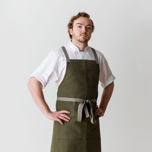 May include: A man wearing a white chef's jacket and a green apron with a grey tie. The apron has a large pocket on the front.