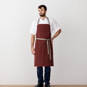 May include: A maroon chef's apron with a brown adjustable strap and a single pocket. The apron is worn by a person standing in front of a white background.