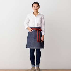 May include: A denim apron with a red ribbon tie. The apron has a single pocket on the front.