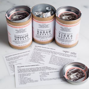 Puede incluir: Tres botes de metal con etiquetas marrones y texto blanco. Las etiquetas dicen "The Reluctant Trading Experiment" y "Bollywood Theater" seguidas de los nombres de diferentes mezclas de especias indias: "Vindaloo Masala", "Caram Masala" y "Tikka Masala". Cada bote tiene una peque&ntilde;a imagen de un coche antiguo en la etiqueta.