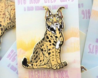 May include: A gold-colored enamel pin featuring an Iberian lynx, a wild cat with a spotted coat. The pin is on a white card with the text "Big and Wild Cats" and "Iberian Lynx" in pink lettering. The card also includes the website "kikidoodle.com".