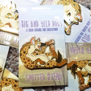 May include: A set of enamel pins featuring spotted hyenas in various poses. The pins are gold-toned and have a glossy finish. The text on the card reads "Big and Wild Dogs: A Hard Enamel Pin Collection" and "Spotted Hyena".