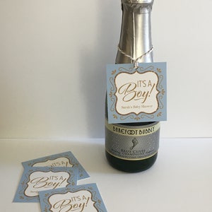 Puede incluir: Una etiqueta de regalo de baby shower azul y dorada con el texto "It's a Boy!" y "Sarah's Baby Shower". La etiqueta est&aacute; unida a una peque&ntilde;a botella de champ&aacute;n.