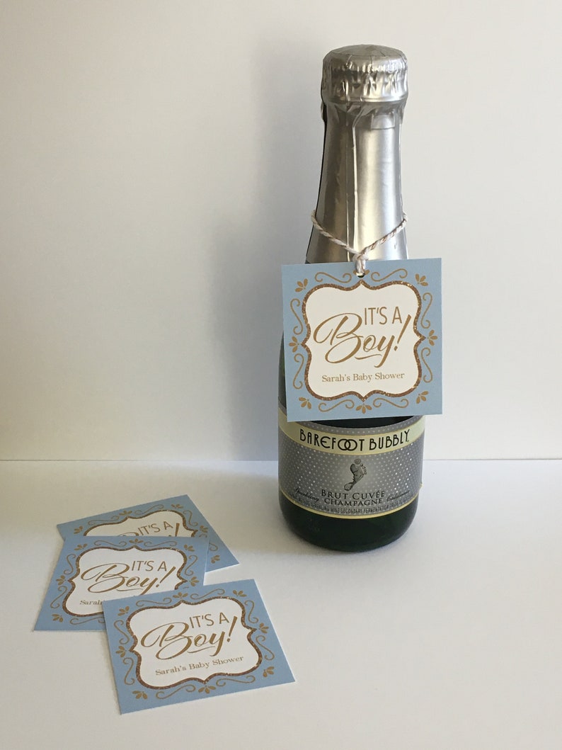 Puede incluir: Una etiqueta de regalo de baby shower azul y dorada con el texto "It's a Boy!" y "Sarah's Baby Shower". La etiqueta est&aacute; unida a una peque&ntilde;a botella de champ&aacute;n.