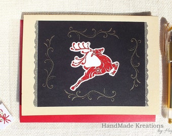 May include: A rectangular greeting card featuring a black center panel with a red reindeer illustration, silver glitter accents, and decorative scrollwork. The card is layered on a red base, accompanied by a small candy cane sticker and a gold pen.