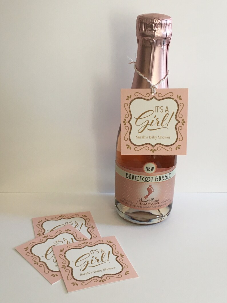 Puede incluir: Etiquetas de regalo de baby shower rosas y doradas 'Es una ni&ntilde;a!' con una botella de champ&aacute;n rosado. La botella est&aacute; etiquetada 'Barefoot Bubbly' y 'Brut Rose Champagne'.