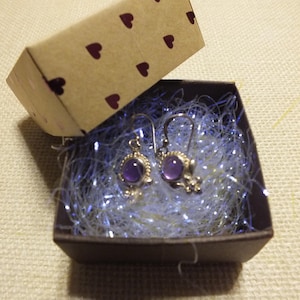 May include: A pair of purple gemstone earrings with silver settings in a small gift box with white tissue paper.