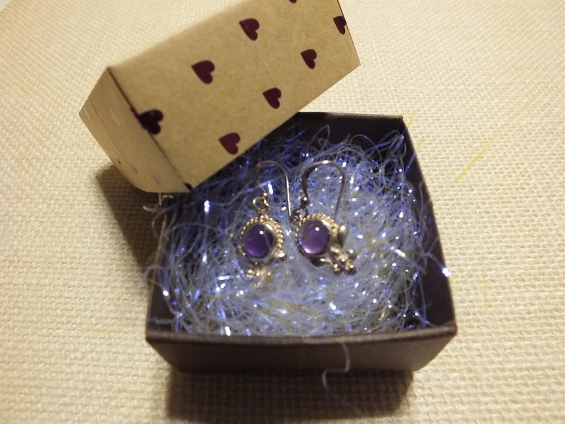 May include: A pair of purple gemstone earrings with silver settings in a small gift box with white tissue paper.