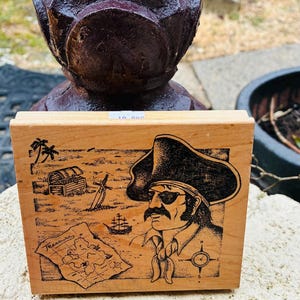 May include: Wooden stamp featuring a pirate with an eye patch, mustache, and hat. The design includes a treasure map, chest, palm tree, and ship. The stamp measures approximately 3 inches in length.