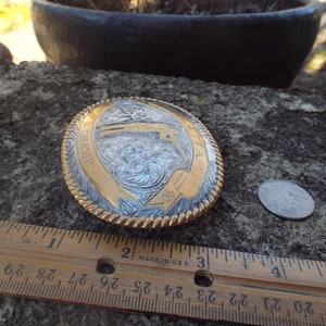 May include: A silver and gold belt buckle with a rope design around the edge. The buckle features a detailed engraving of a cowboy riding a horse with the word "CHAMP" and the number "90" on it.