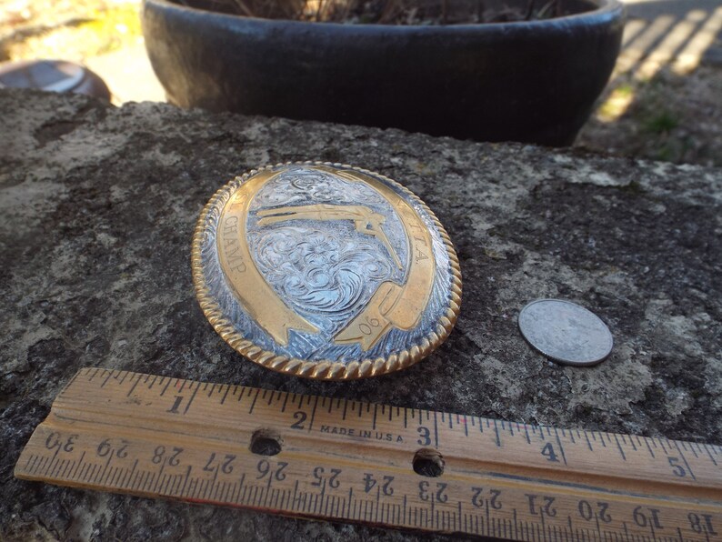 May include: A silver and gold belt buckle with a rope design around the edge. The buckle features a detailed engraving of a cowboy riding a horse with the word "CHAMP" and the number "90" on it.