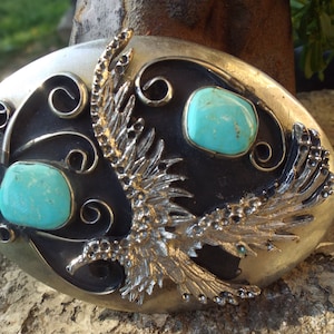 May include: Oval silver-tone belt buckle with a black center, featuring two turquoise stones and a detailed silver-tone bird design. The buckle has a decorative, ornate style.
