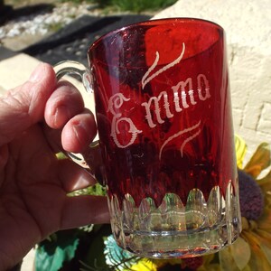 May include: A red glass mug with a clear handle. The mug has a scalloped rim and is etched with the name "Emma".