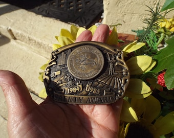 May include: A gold-toned metal belt buckle with an engraved design featuring a city skyline, a cornucopia, and a windmill. The buckle has the text "City of East Moline, Illinois" and "1984-1985 Safety Award".