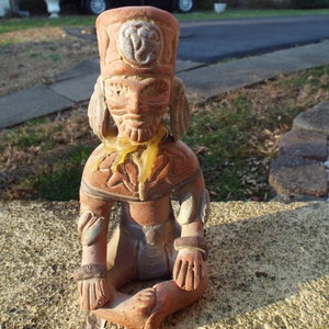 May include: A terracotta figurine of a seated figure with a headdress and a wax drip down the front. The figurine is wearing a decorative garment with a floral pattern.