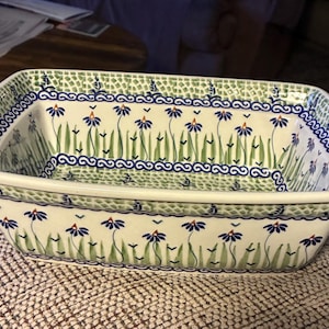 May include: A rectangular ceramic baking dish with a cream-colored base, featuring a hand-painted pattern of blue flowers with green stems and leaves. The rim is decorated with a blue swirling border and a green mosaic-style textured edge.