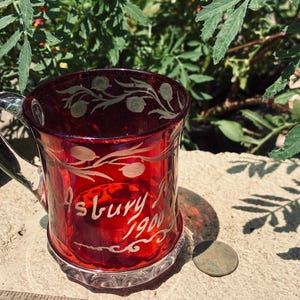 May include: A ruby red glass mug with a clear glass handle, featuring etched floral vines and the text "Asbury Park 1900". A wooden ruler is positioned at the base, showing the mug is approximately 3 inches wide at the top rim.