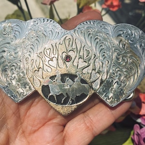 May include: A heart-shaped silver belt buckle featuring intricate scrollwork engravings, a central gold-toned flame motif with a small embedded red gemstone, and a black oval inlay depicting two figures riding horses in profile.