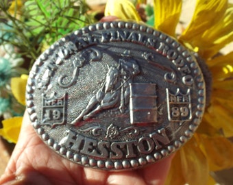 May include: Silver belt buckle with a rodeo scene featuring a horse and barrel racer. The buckle is oval shaped and has the text "National Finals Rodeo" and "Hesston" on it. The buckle also has the text "NFR 19" and "NFR 89" on it.