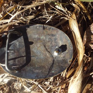 May include: A silver-toned belt buckle with a worn finish. The buckle has a rounded shape and a simple design. The back of the buckle has the words "AUSTRIA" and "SILVER" engraved on it.