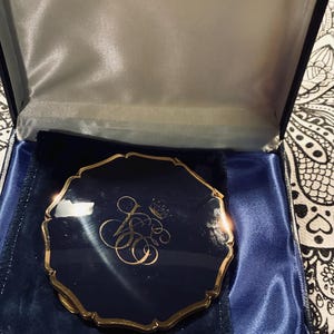 May include: A round, dark navy blue compact mirror with a scalloped gold-tone metal edge. The lid features a gold-colored monogram of the letters J and S topped with a small crown. The compact is presented inside a dark blue velvet-lined gift box.