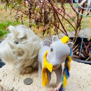 May include: Two toy ponies are displayed outdoors. One is a fluffy white pony with long hair. The other is a gray pony with a yellow scarf and a multicolored braid. A coin is visible in the foreground.