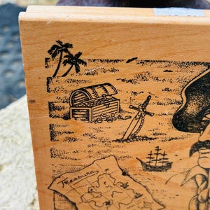 May include: Wooden block with a pirate-themed illustration. The design includes a treasure chest, map, sword, palm trees, and a ship. The word "Treasure" is written on the map. The block is light brown.