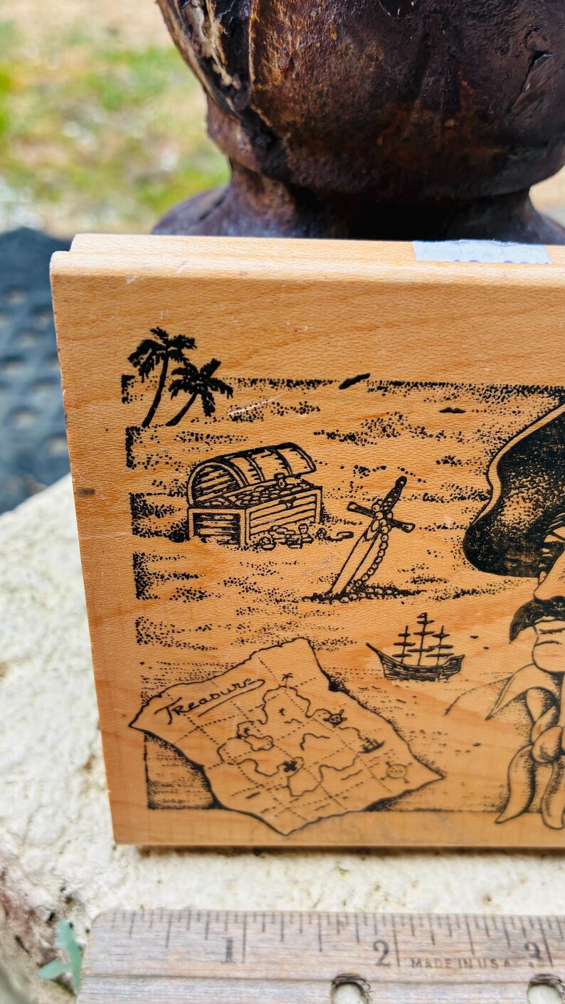May include: Wooden block with a pirate-themed illustration. The design includes a treasure chest, map, sword, palm trees, and a ship. The word "Treasure" is written on the map. The block is light brown.