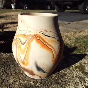 May include: A small, white ceramic vase with a marbled design in shades of orange, brown, and black. The vase has a rounded body and a slightly flared lip.