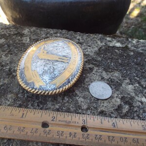 May include: A silver and gold belt buckle with an engraved design of a person holding a rifle. The buckle is oval shaped and has the text "SL CHAMP" on it. The buckle is about 2 inches in diameter.