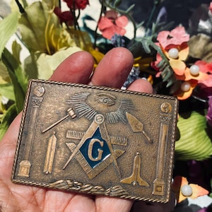 May include: A rectangular bronze-toned metal belt buckle featuring embossed Masonic symbols including a central square and compass with a blue enamel inlay letter G, an eye, a gavel, a trowel, and two pillars, all framed by a rope-style border.