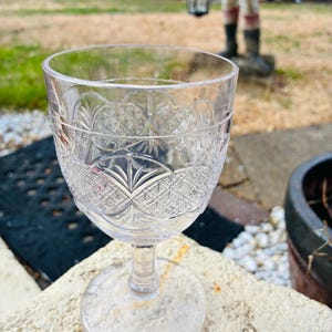 May include: A clear, cut-glass wine glass with a detailed pattern on the bowl. The glass sits on a small, round base and has a slender stem. A silver coin rests nearby. The background is blurred, showing outdoor elements.