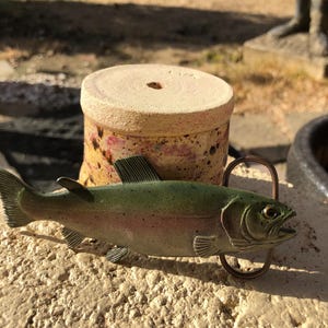 May include: A decorative fish-shaped belt buckle with a bronze-colored metal loop. The fish is painted in shades of green, pink, and silver. A ceramic pot with a hole in the center is in the background.
