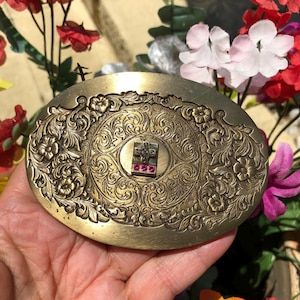 May include: Oval-shaped brass belt buckle with intricate floral and scrollwork design. The center features a small square with a cross and three red gemstones. The buckle is held in a hand, surrounded by colorful flowers.